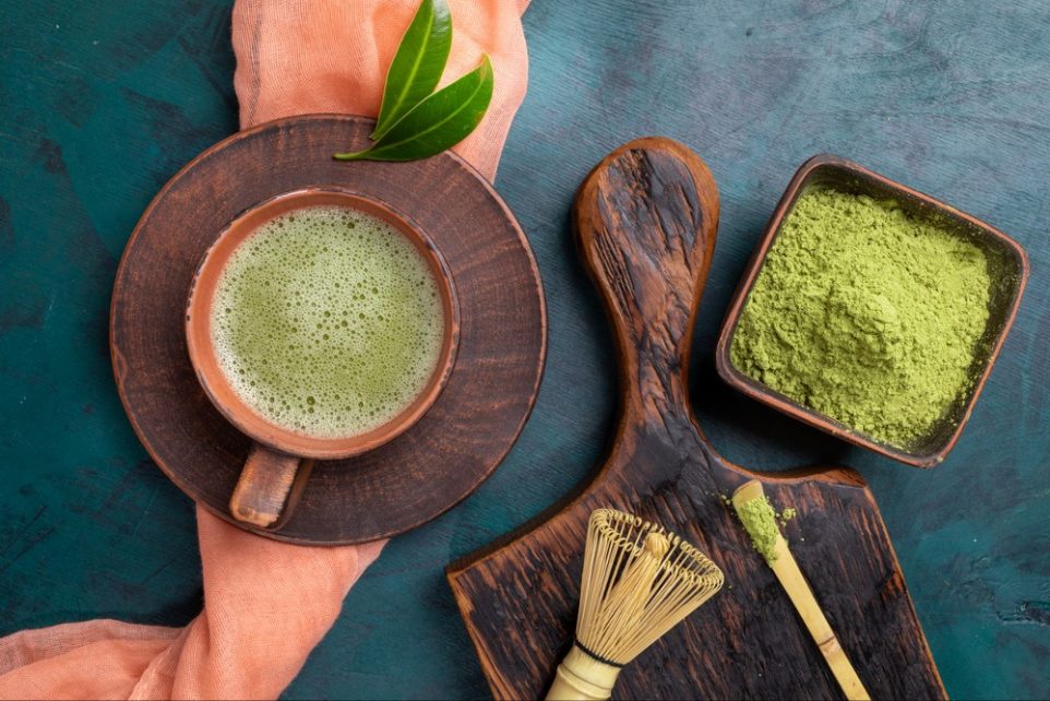 Green matcha tea and powder in brown cups and whisk and spoon on wooden serving board on emerald backdrop with coral napkin